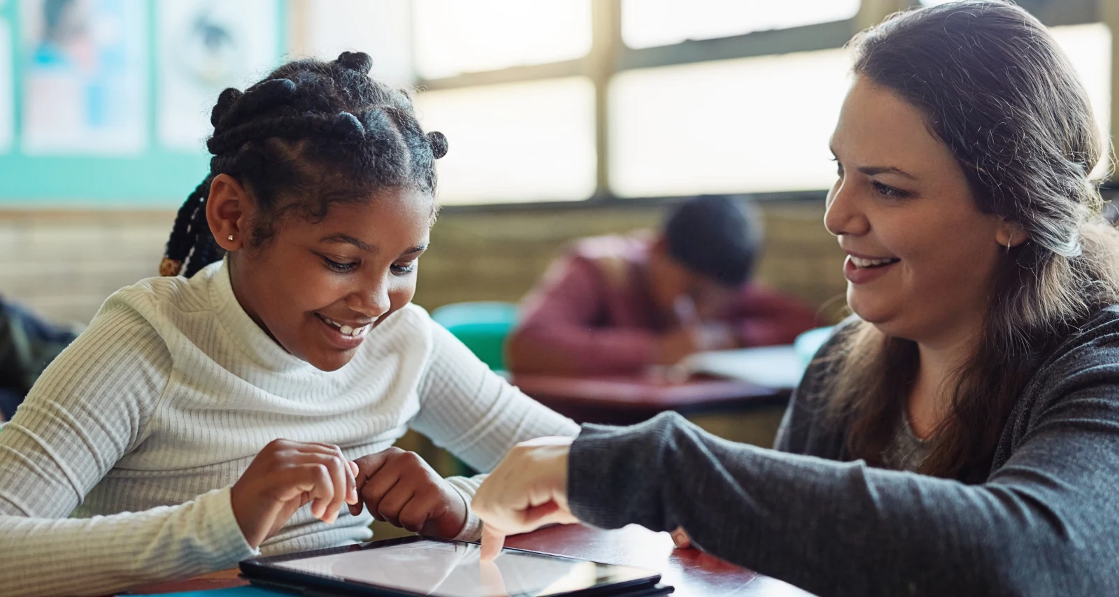 A smiling student plays a Playbl game on a tablet as her teacher points to the screen